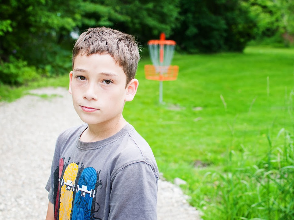 a boy stands on Farmington's Disco Golf course with a pathway and green grass behind him and the disc golf game. 