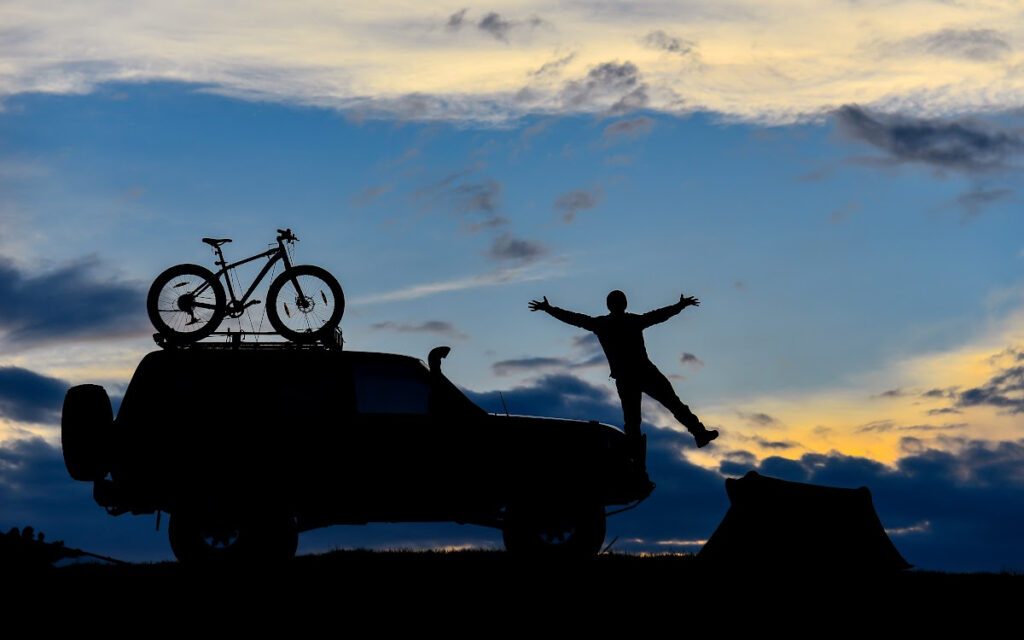 A man jumps joyfully into a sunset near his S.U.V. with his bicycle strapped to the top. 