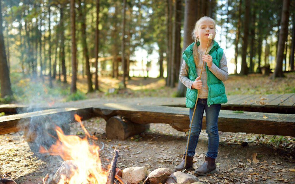 A blonde little girl in a green vest toasts marshmallows at a campfire.